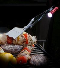 BBQ Croc 21-inch barbecue tongs with flashlight holding a chicken skewer over a grill full of food illuminated by the clip-on light.