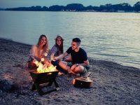 Three young people sitting on a beach around a BBQ Croc EASY GRILL, the upper grill of the barbecue is removed and they are grilling marshmallows over the open flame, they have their backs to the water in the background.