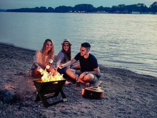 Three young people sitting on a beach around a BBQ Croc EASY GRILL, the upper grill of the barbecue is removed and they are grilling marshmallows over the open flame, they have their backs to the water in the background.