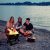 Three young people sitting on a beach around a BBQ Croc EASY GRILL, the upper grill of the barbecue is removed and they are grilling marshmallows over the open flame, they have their backs to the water in the background.